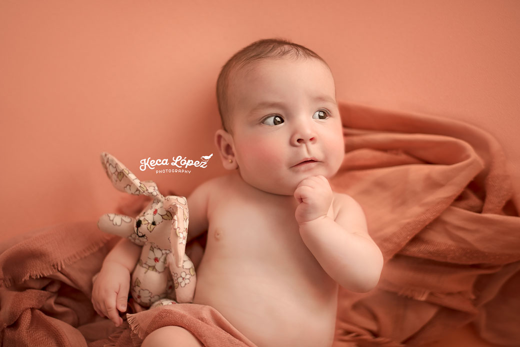 Bonita bebé abrazando un peluche de conejo sobre puff color salmón. Tiene una posición pensativa