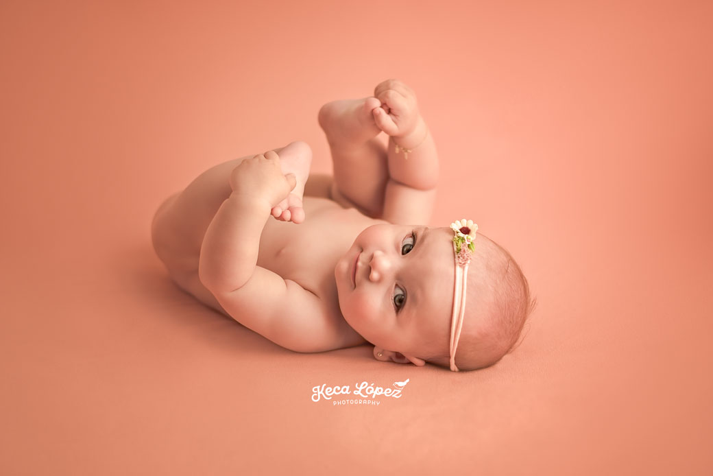 Bebé niña de 4 meses tocandose los pies, sonriendo y con una dulce mirada. Fondo de color rosita. Lleva una diadema en la cabeza con una flor