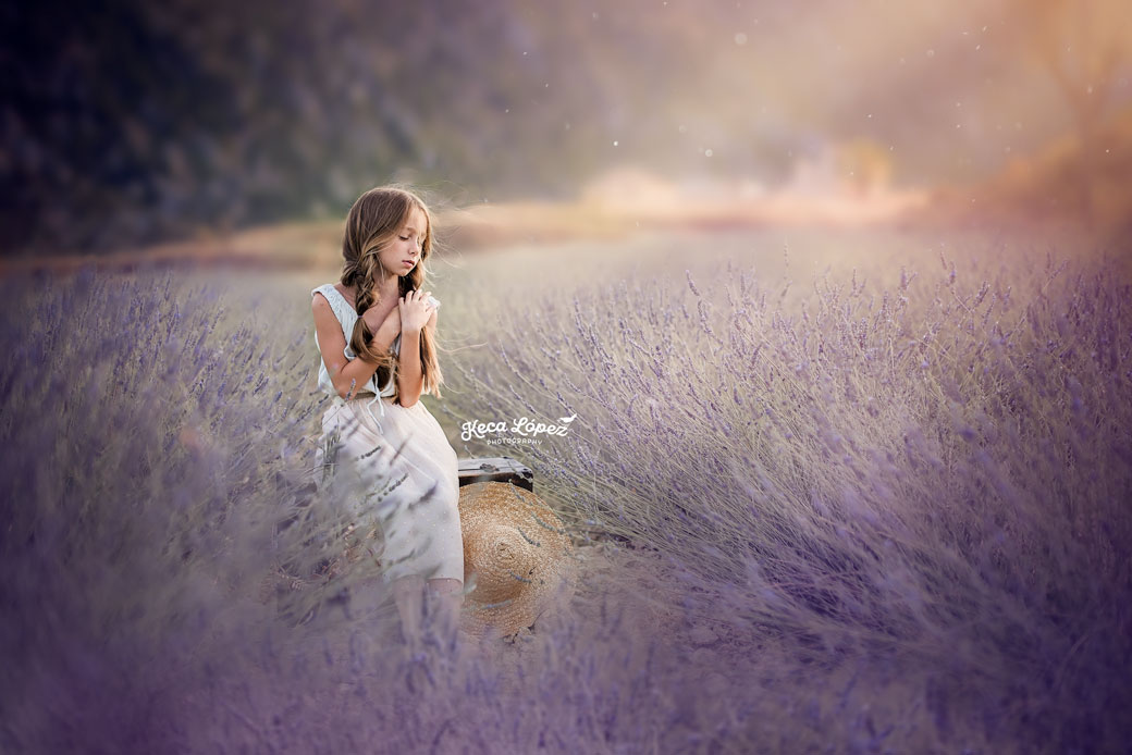 Niña sentada en un campo de lavanda Campo de San Juan. Lleva un vestido blanco. En sus pies tiene un sombrero de paja