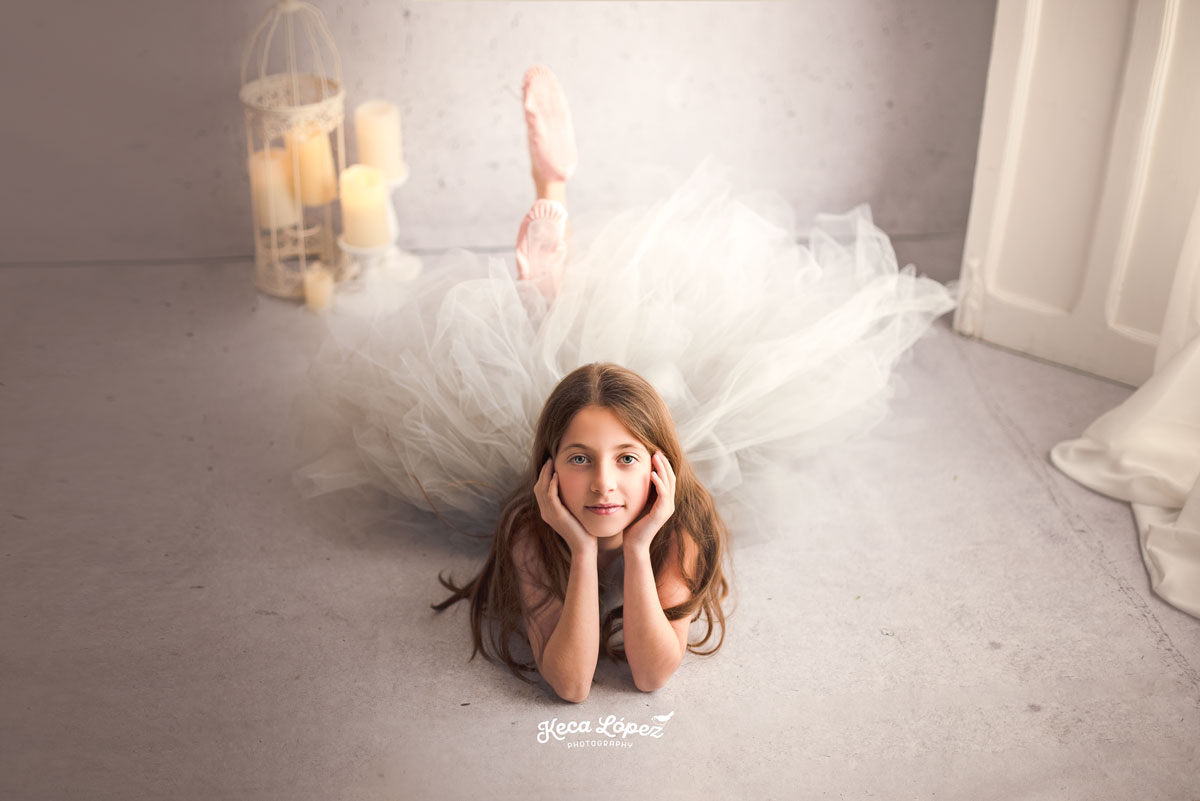 Bailarina en el suelo, su cabeza reposa sobre sus manos. Su vestido es de tull de color gris y el escenario es blanco.