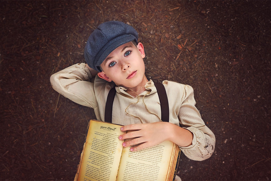 niño en el suelo con gorro azul tirantes y un libro